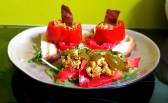 Großaufnahme; 2 gefüllte rote Paprikaringe auf getoastetem Weißbrot angerichtet auf einem weißen Teller; In der Fülle steckt jeweils eine kleine Scheibe angebratener veganer Speck; Im Vordergrund als Beilage Vogerlsalat mit rotem Paprika etwas Rührei und Chalapenós. Der Teller ist mit kleinen Salatstückchen garniert.