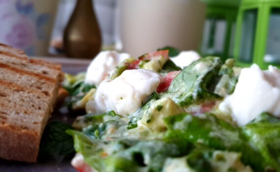 Nahansicht, links ein getoastetes Schwarzbrot mit Toaststreifen; rechts der auf einen grauen Teller drapierte Salat; Zerrissene Salatblätter, ganze Basilikumblätter, Tomaten- und Mozzarellastücke sind mit sämigem Pesto angerichtet. Es sieht sehr saftig aus; Ein paar weiße Mozzarellastücke liegen oben auf und glänzen im Licht.