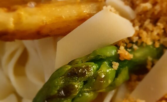 Auf gekochten gelblich weißen Bandnudeln liegen angebratene grüne und weiße Sparkel; darauf die gerösteten Semmelbrösel; Die Speise ist mit Parmesanstücken garniert. Im Vordergrund ist die Spitze eines grünen Spargels zu sehen der leicht bräunliche Brat-Flecken aufweist und noch vom Öl in der Pfanne glänzt. Auf der Spargelspitze liegen ein Parmesanstück und ein paar Brösel. Der Hintergrund mit weiteren weißen und grünen angebratenen Spargeln, Nudeln und Brösel verläuft in Unschärfe; Aufnahme von schräg oben auf den grauen Teller von dem man nur einen kleinen Teil noch im Hintergrund erkennen kann; Dahinter liegt eine zum Dreieck gefaltene Serviette.