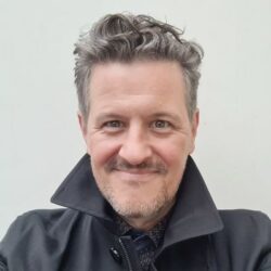 Weißer Mann in schwarzer Jacke mit hochgestelltem Kragen, lockiges kurzes Haar und 3-Tages-Bart; lächelt freundlich.