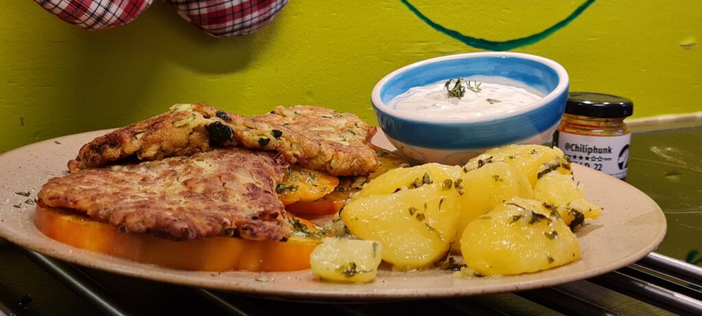 Teller mit 2 Zucchinischnitzel auf großen Tomatenscheiben (Ochsenherz); daneben glänzende Petersilkartoffel; 1 kleine Schale mit Sauce Tartare und Kräutertopping; 1 Glas Chiliphunk Chilipaste, neben dem Teller im Hintergrund.