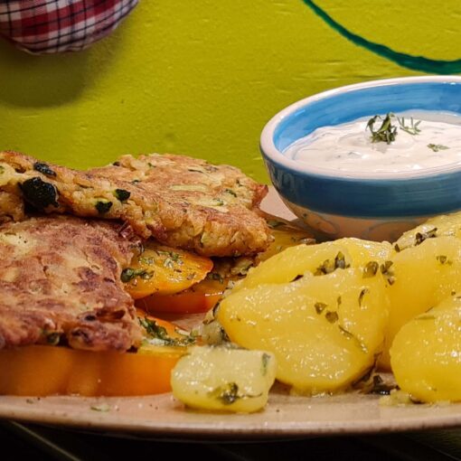 Teller mit 2 Zucchinischnitzel auf großen Tomatenscheiben (Ochsenherz); daneben glänzende Petersilkartoffel; 1 kleine Schale mit Sauce Tartare und Kräutertopping; 1 Glas Chiliphunk Chilipaste, neben dem Teller im Hintergrund.