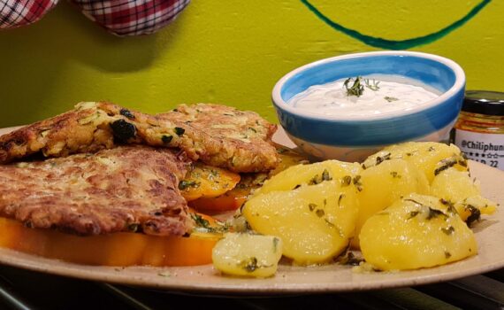Teller mit 2 Zucchinischnitzel auf großen Tomatenscheiben (Ochsenherz); daneben glänzende Petersilkartoffel; 1 kleine Schale mit Sauce Tartare und Kräutertopping; 1 Glas Chiliphunk Chilipaste, neben dem Teller im Hintergrund.