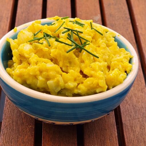 Kleine Keramikschüssel mit gelbem Aufstrich. Oben auf ein paar Schnittlauchteilchen. Die Schüssel ist weiß-blau mit Ornamenten verziert. Sie steht auf einem Holztisch.