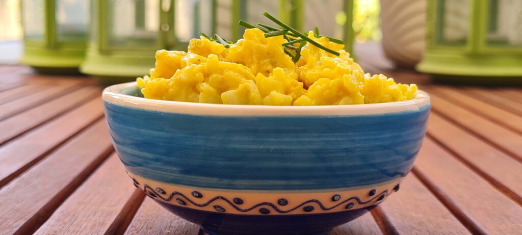 Kleine Keramikschüssel mit gelbem Aufstrich. Oben auf ein paar Schnittlauchteilchen. Die Schüssel ist weiß-blau mit Ornamenten verziert. Sie steht auf einem Holztisch.