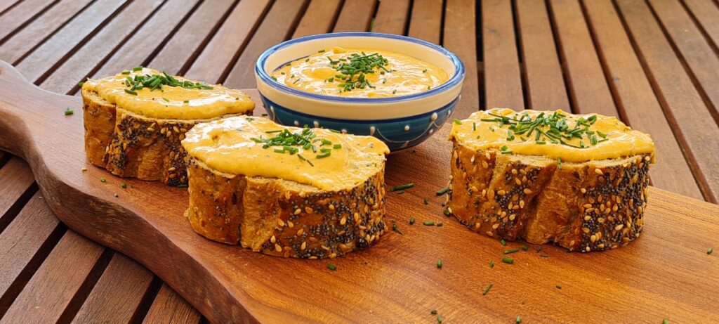 Drei Scheiben vom Mehrkorn-Baguette mit Liptauer, vor einer kleinen blau-weiß verzierten Schale mit Liptauer; alles mit Schnittlauch-Topping; Angerichtet auf einem geschwungenen Edelholzbrett, auf einem Garten-Holztisch.