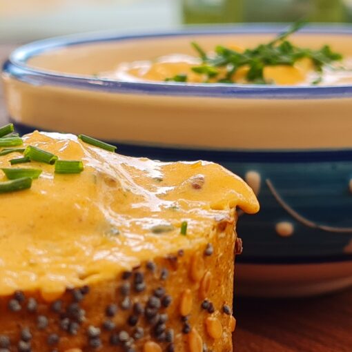 Nahaufnahme; Zwei Scheiben vom Mehrkorn-Baguette mit Liptauer, vor einer kleinen blau-weiß verzierten Schale mit Liptauer; alles mit Schnittlauch-Topping; Angerichtet auf einem Edelholzbrett, auf einem Garten-Holztisch.