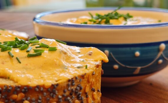 Nahaufnahme; Zwei Scheiben vom Mehrkorn-Baguette mit Liptauer, vor einer kleinen blau-weiß verzierten Schale mit Liptauer; alles mit Schnittlauch-Topping; Angerichtet auf einem Edelholzbrett, auf einem Garten-Holztisch.