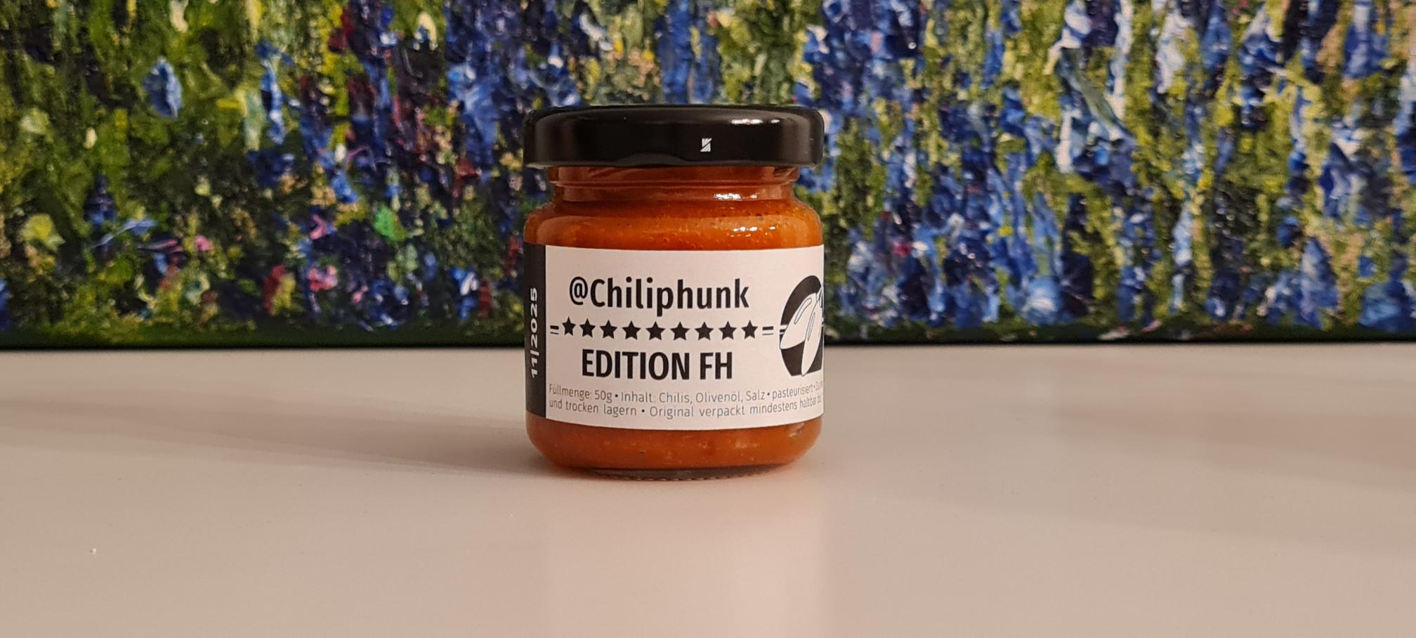 Ein Glas Chiliphunk "Edition FH" mit 9 Sternen, vor einem Gemälde einer Blumenwiese.
