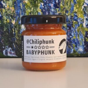 1 Glas Chilipaste Chiliphunk "Babyphunk" mit 3 Sternen am Etikett. Im Hintergrund steht ein Ölgemlde, von dem man nur den Fußbereich sieht. Grün-blaue Struktur mit lila Punkten.