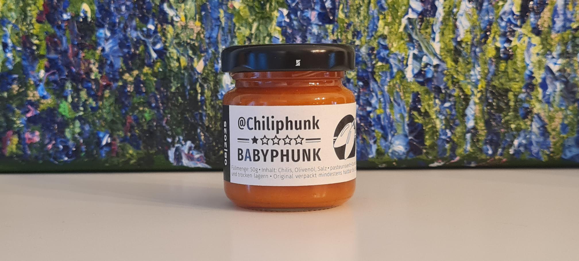 1 Glas Chilipaste Chiliphunk "Babyphunk" mit 3 Sternen am Etikett. Im Hintergrund steht ein Ölgemlde, von dem man nur den Fußbereich sieht. Grün-blaue Struktur mit lila Punkten.
