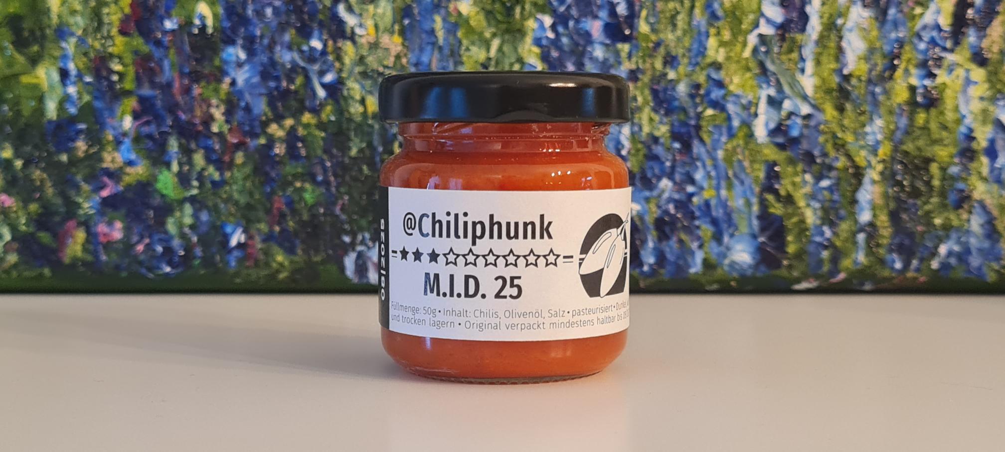 1 Glas Chilipaste Chiliphunk "M.I.D. 25" mit 3 Sternen am Etikett. Im Hintergrund steht ein Ölgemlde, von dem man nur den Fußbereich sieht. Grün-blaue Struktur mit lila Punkten.